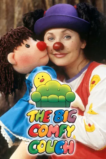 The poster features a woman in a purple hat and red nose, holding a plush doll with a yellow duck on its head. The background is warm and cozy, with soft lighting. The title "The Big Comfy Couch" is displayed in colorful, playful letters. The overall mood is cheerful and inviting, suggesting a re...