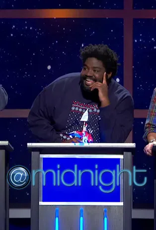 A comedian stands behind a podium at "Midnight with Chris Hardwick," laughing heartily while wearing a dark sweater with a colorful graphic. The starry backdrop suggests a night sky, and the cool blue tones of the set enhance the nighttime theme.