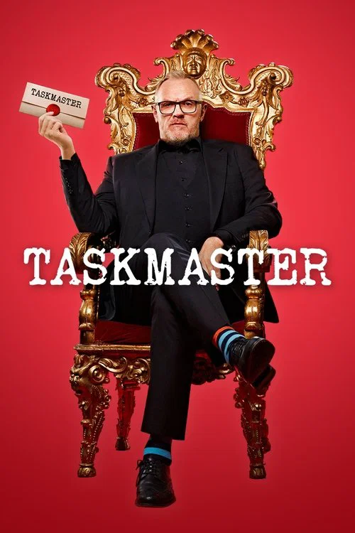 A man in a black suit sits atop an ornate golden throne against a vibrant red background, holding a card that reads "TASKMASTER." The dramatic lighting and bold colors evoke a sense of authority and challenge, capturing the essence of the show's humorous and competitive spirit.