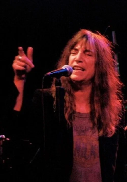 A passionate woman with long hair performs on stage, her vibrant red lighting illuminates her expressive face and hair against a dark background.